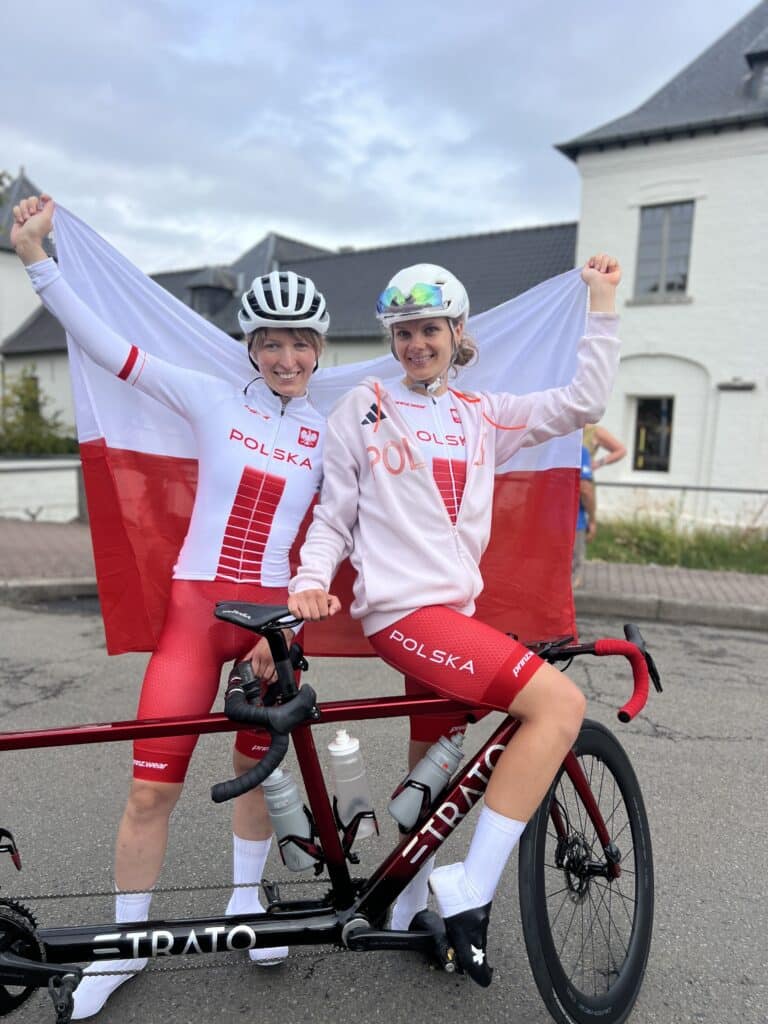 Patrycja Kuter i Karolina Kołkowicz pozują do zdjęcia przy swoim tandemie. Obie są w strojach startowych reprezentacji Polski, za plecami trzymają polską flagę.