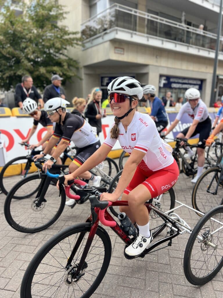 Rita Malinkiewicz w stroju reprezentacji Polski siedzi na trójkołowym rowerze na starcie wyścigu. W tle inne zawodniczki