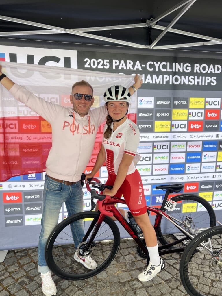 Rita Malinkiewicz pozuje do zdjęcia w stroju reprezentacji Polski i na trójkołowym rowerze, obok niej trener z flagą rozpostartą za plecami
