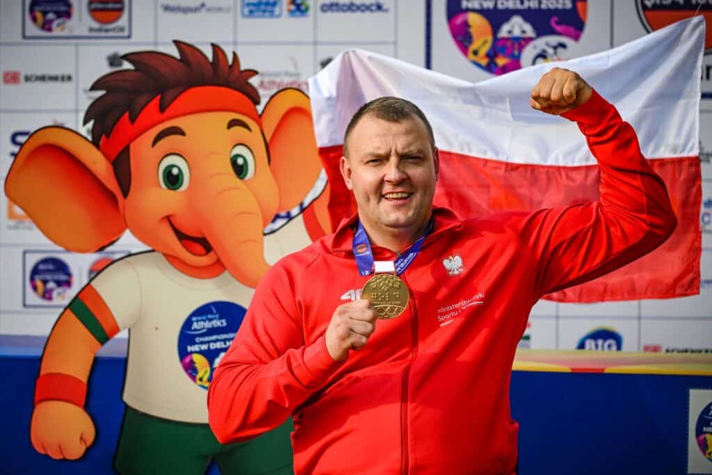 Bartosz Górczak prezentuje swój złoty medal, napinając biceps. Za nim polska flaga i słonik, czyli maskotka mistrzostw świata