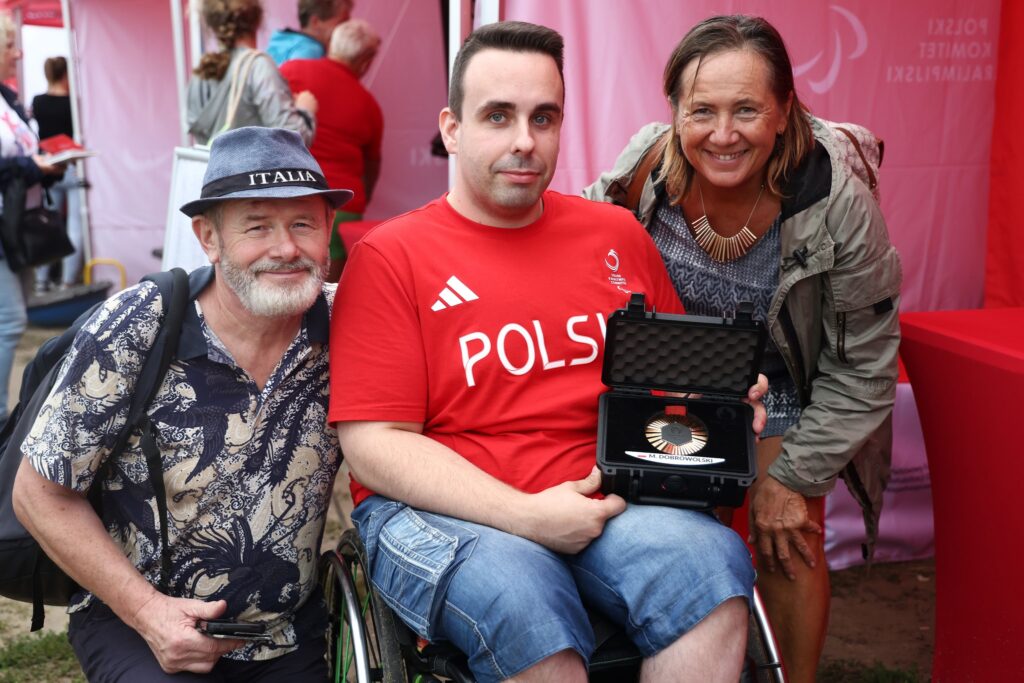 Siedzący na wózku Marek Dobrowolski prezentuje swój brązowy medal igrzysk z Paryża. Obok niego do zdjęcia pozują kobieta i mężczyzna.