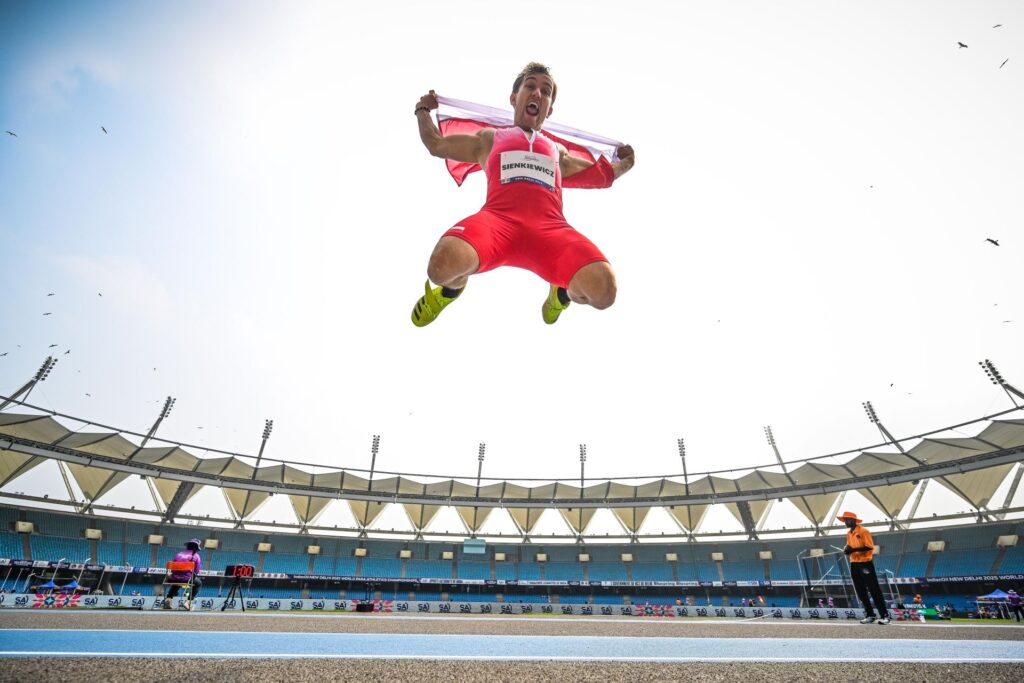 Radosny Bartosz Sienkiewicz skacze bardzo wysoko w górę z polską flagą za plecami. W tle trybuny stadionu