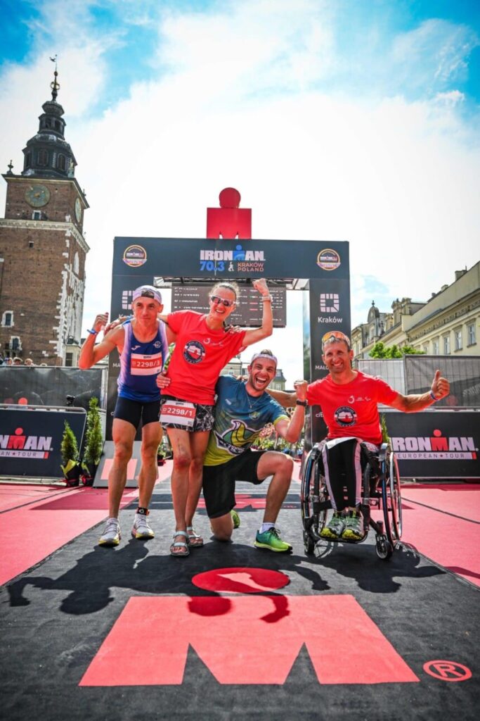 Cztery osoby, w tym jedna na wózku, pozuje do zdjęcia na mecie zawodów IronMan