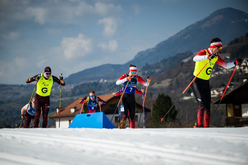 Grupa zawodniczek z przewodnikami na trasie biegu narciarskiego. Na pierwszym planie Aneta Kobryń z przewodnikiem Bartłomiejem Puto. W tle góry