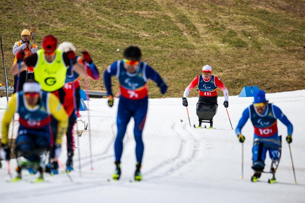 Narciarze biegowi na zaśnieżonej trasie. Z przodu kilku zawodników stojących lub jadących na siedząco. W pewnej odległości za nimi Krzysztof Plewa