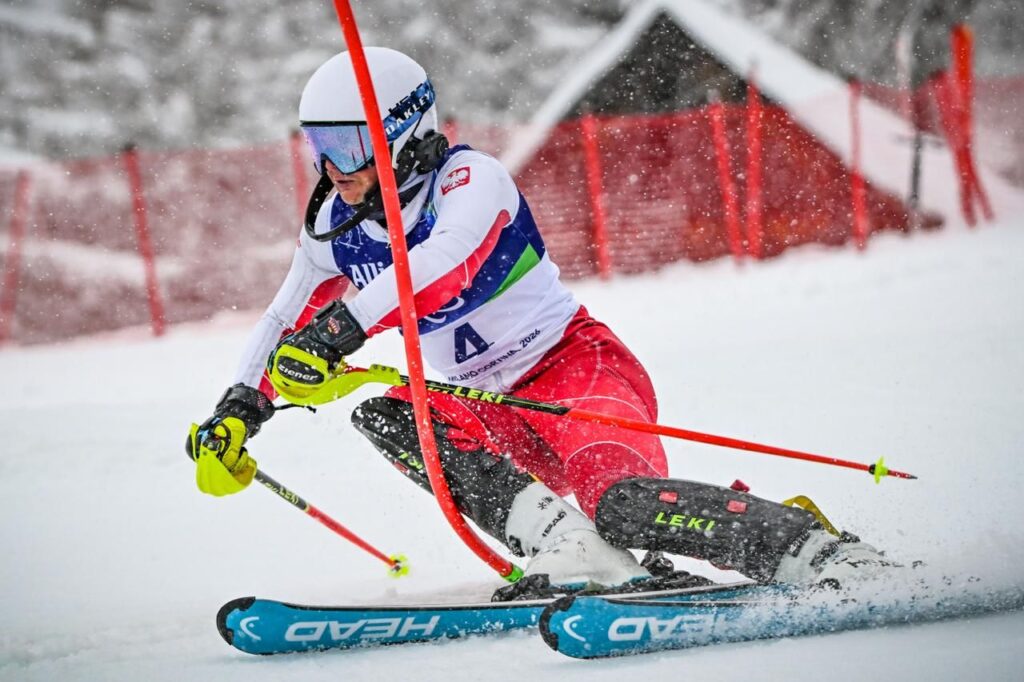 Michał Gołaś jadąc po śniegu na nartach w dynamicznym skręcie mija tyczkę slalomową, która ugina się pod jego naporem