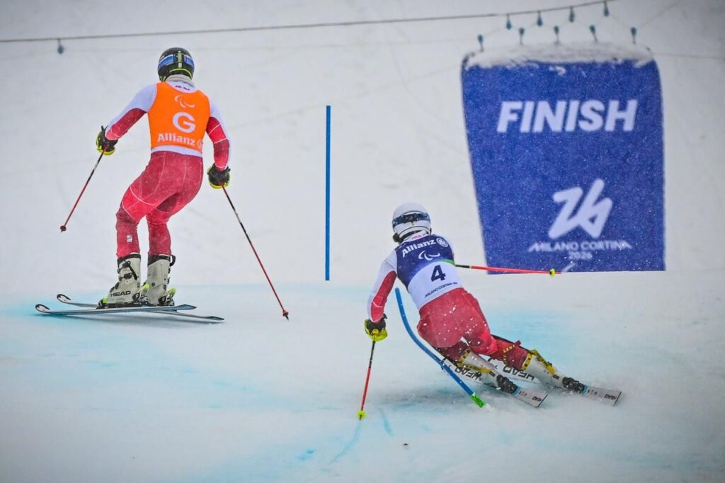 Michał Gołaś jedzie po śniegu na nartach, mijają tyczkę slalomową, za swoim przewodnikiem Kacprem Walasem. W tle widać napis: finish
