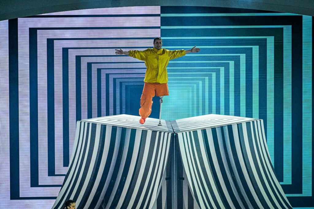 Scena ceremonii otwarcia. Na czymś w rodzaju podestu stoi mężczyzna, który zamiast jednej nogi ma protezę